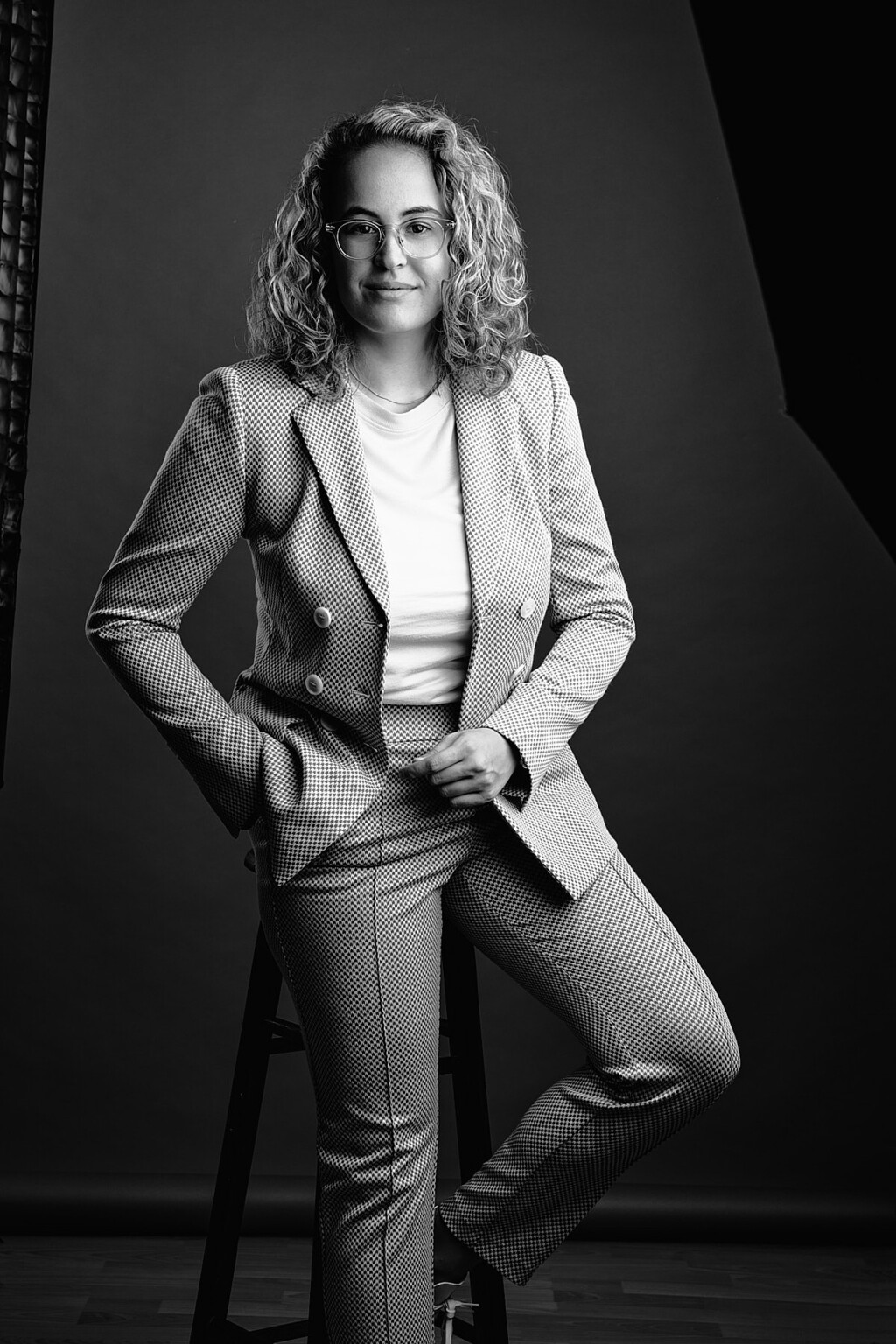 Schwarzweiß Portrait einer Frau mit Locken im hellen Anzug auf einem Hocker