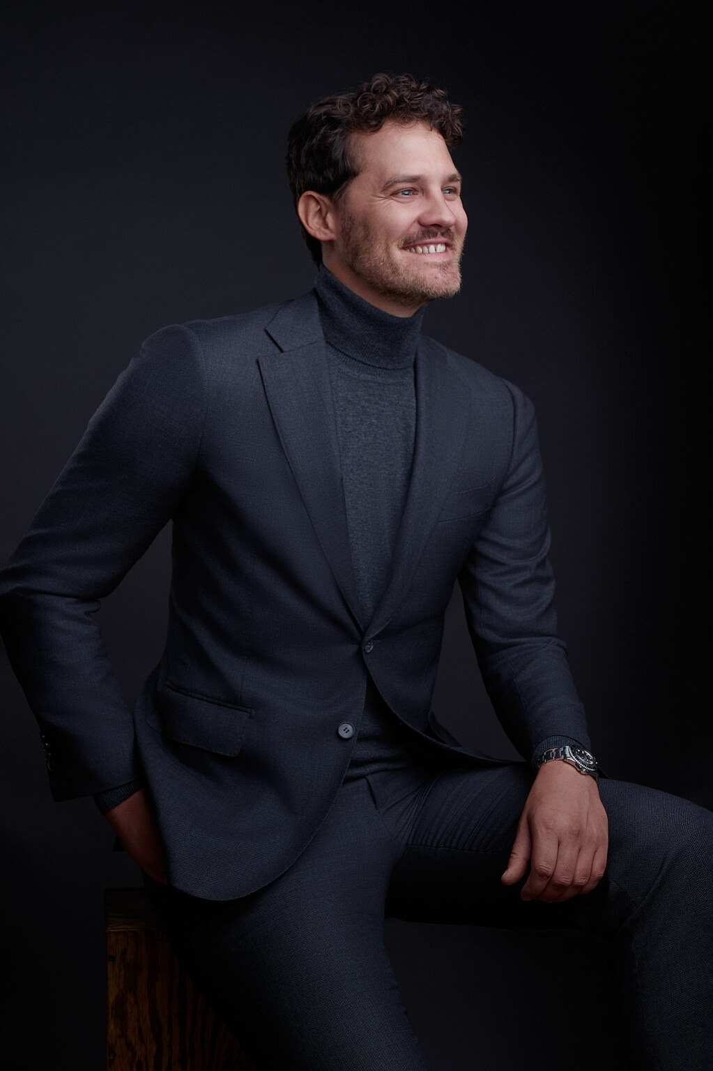 Sitzendes Business Portrait: Mann in Anzug und Rollkragen lächelt vor schwarzem Hintergrund, Frankfurt