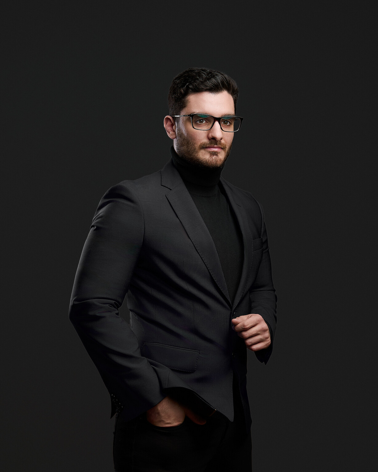 Mann mit Brille in dunklem Jackett und Rollkragen vor dunklem Hintergrund, Mitarbeiterportrait