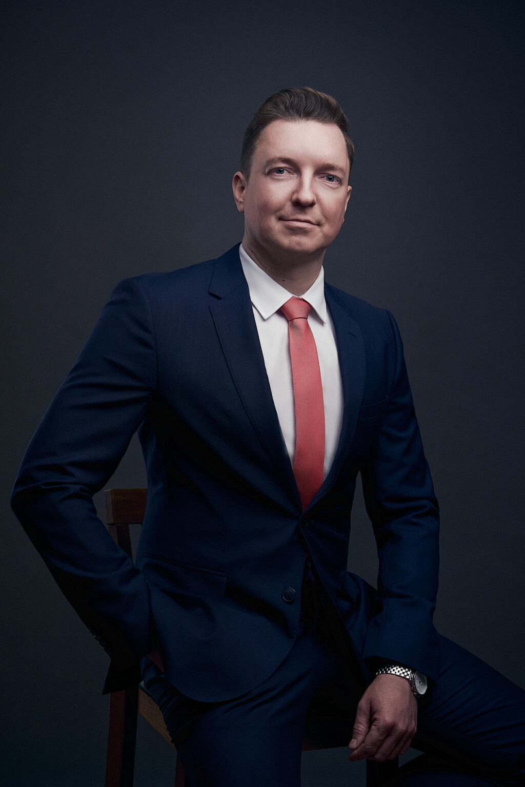 Mann in dunkelblauem Anzug mit roter Krawatte sitzt vor dunklem Hintergrund, Executive Portrait