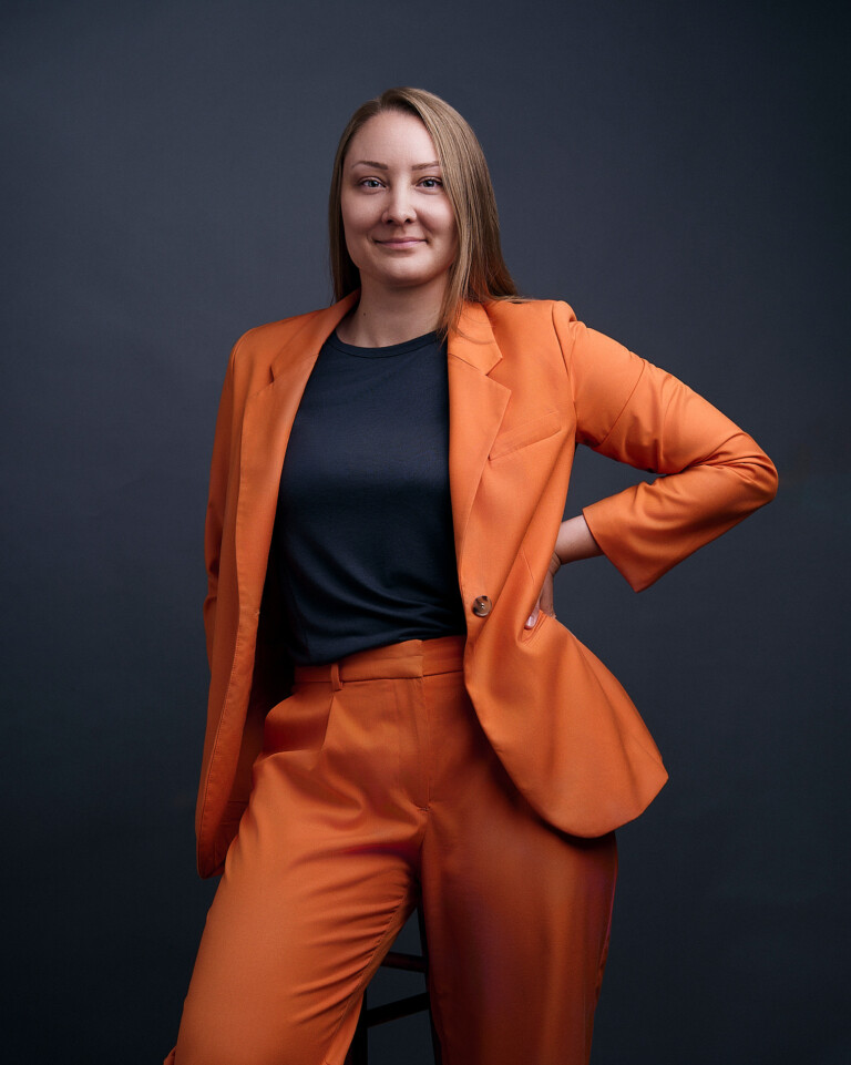 Frau im orangefarbenen Anzug mit einer Hand an der Hüfte vor dunklem Hintergrund