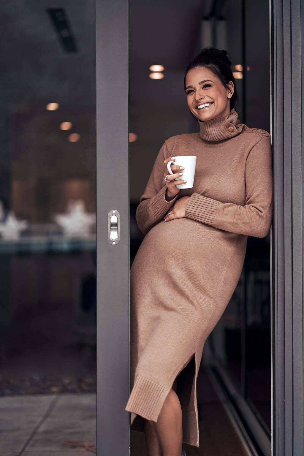 Amira Aly beim Babybauch-Shooting im Strickkleid mit Tasse an der Tür