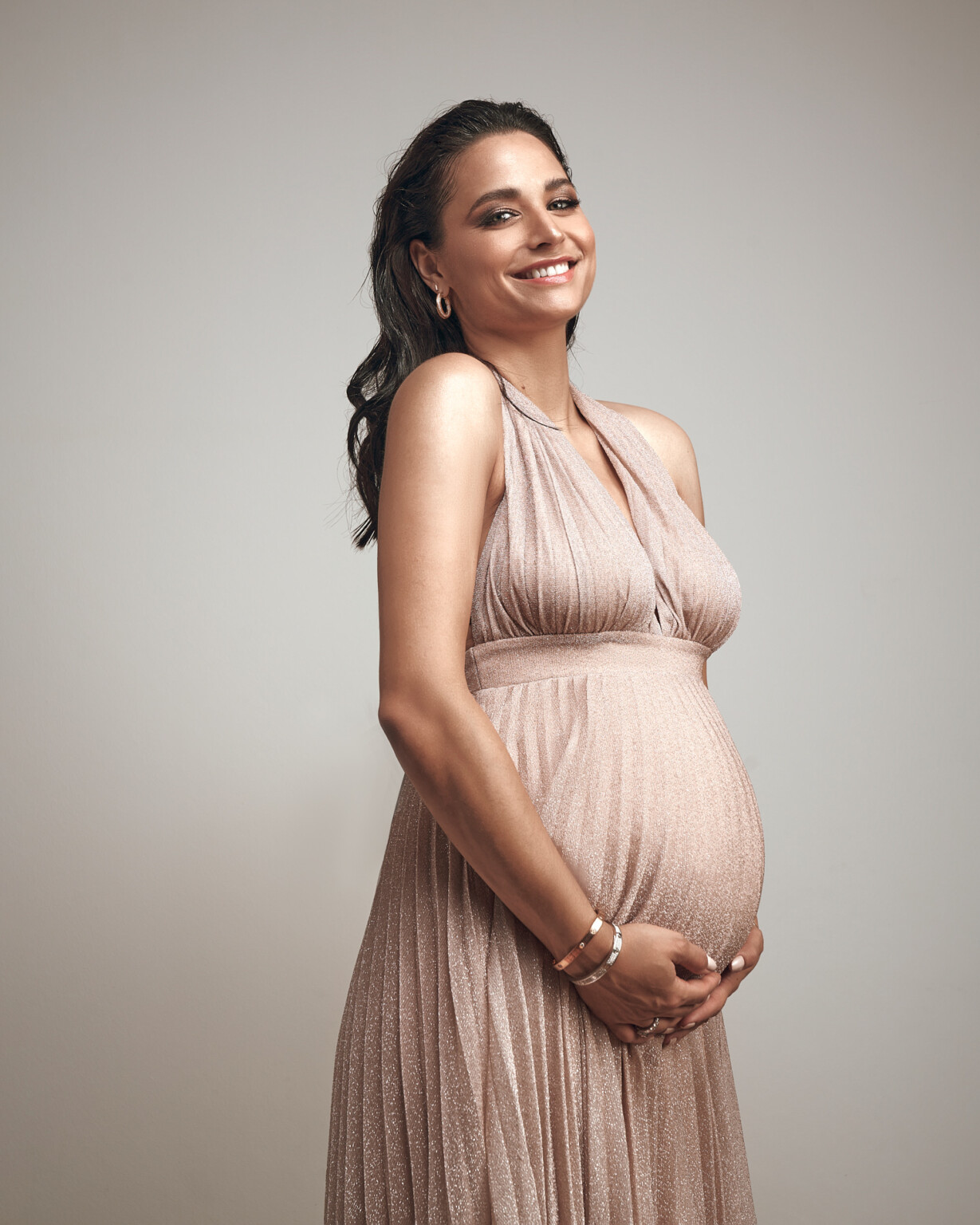 Amira Aly lächelnd im plissierten Kleid in Rosé, Hände am Babybauch