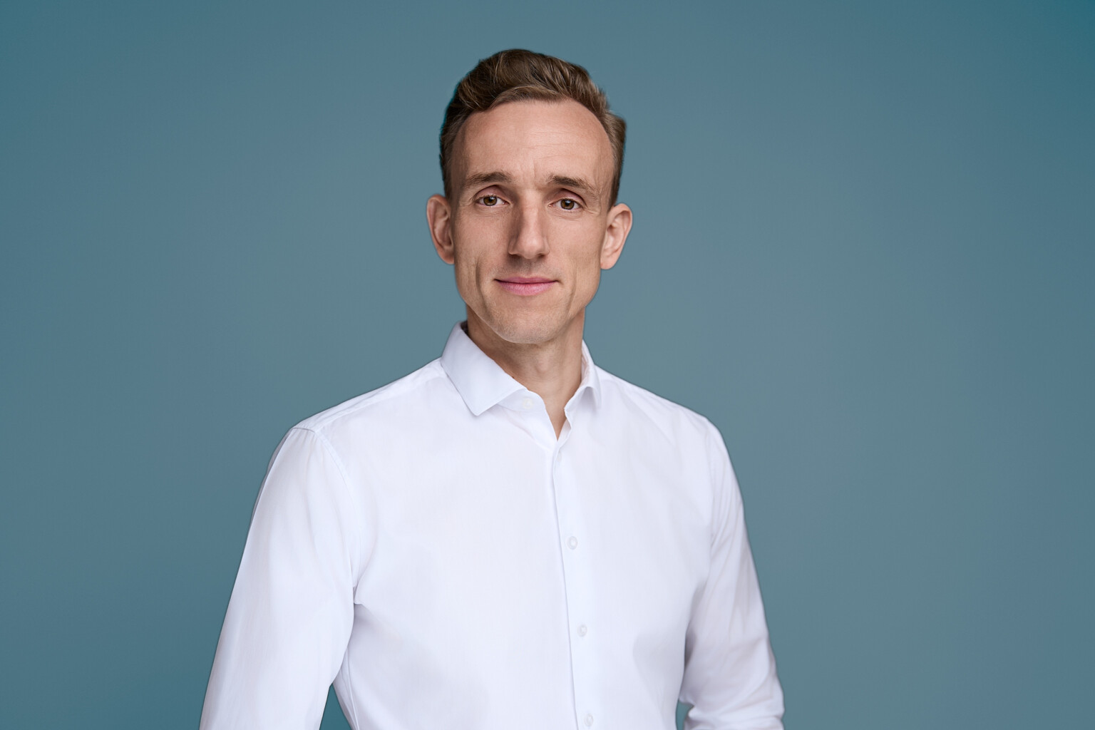 Business-Casual-Portrait eines Mannes im weißen Hemd ohne Sakko vor petrolblauem Studiohintergrund
