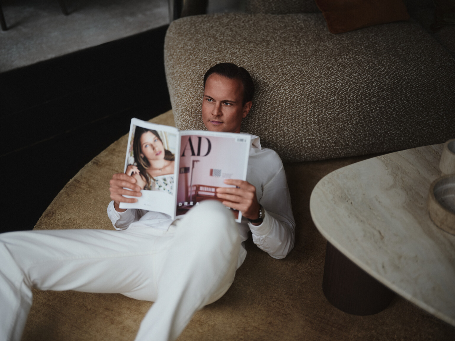 Mann in weißem Hemd und weißer Hose liest entspannt ein Magazin auf einem Bouclé-Sofa