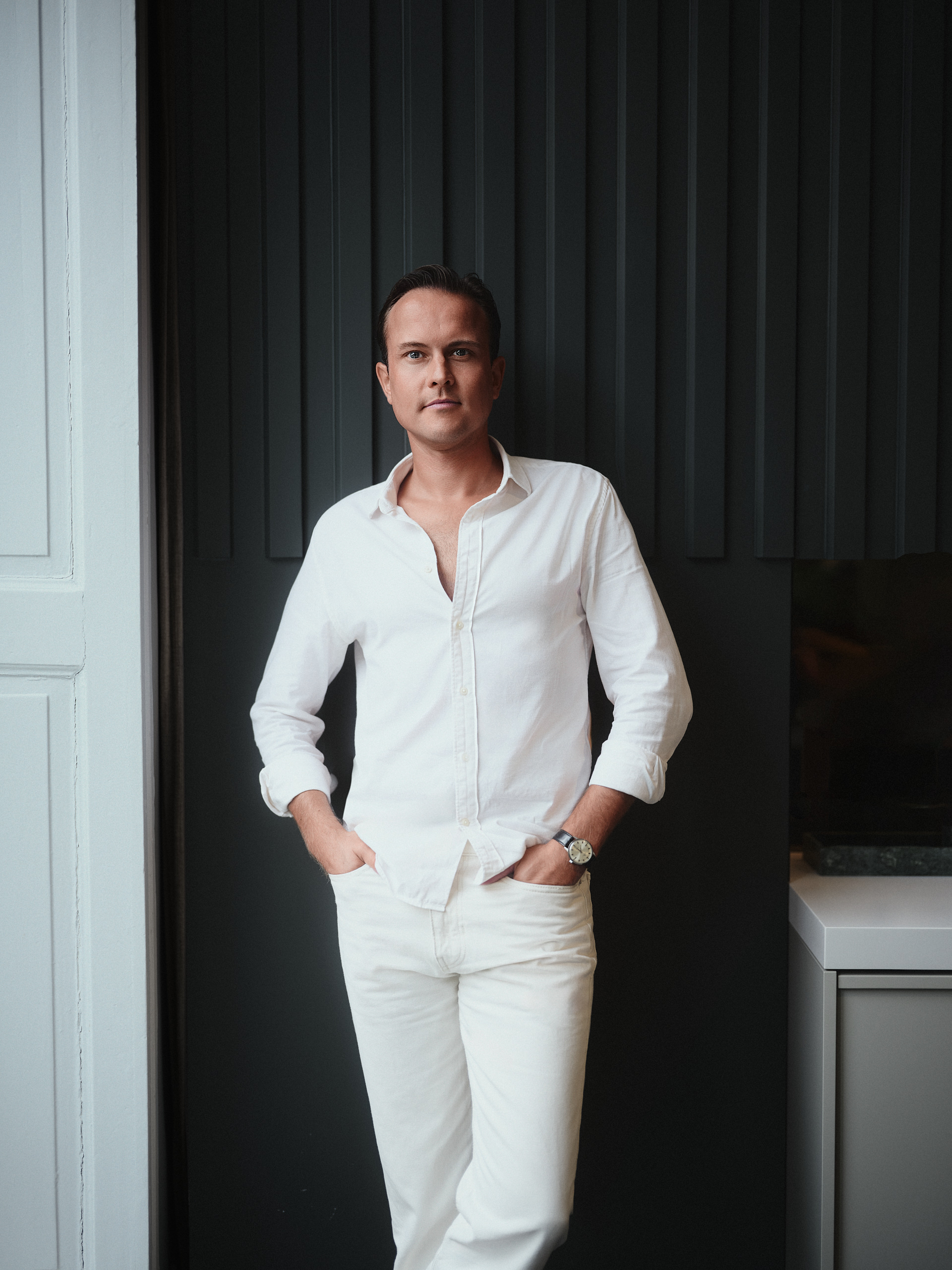 Man dressed all in white standing casually in front of a dark slatted wall, hands in pockets