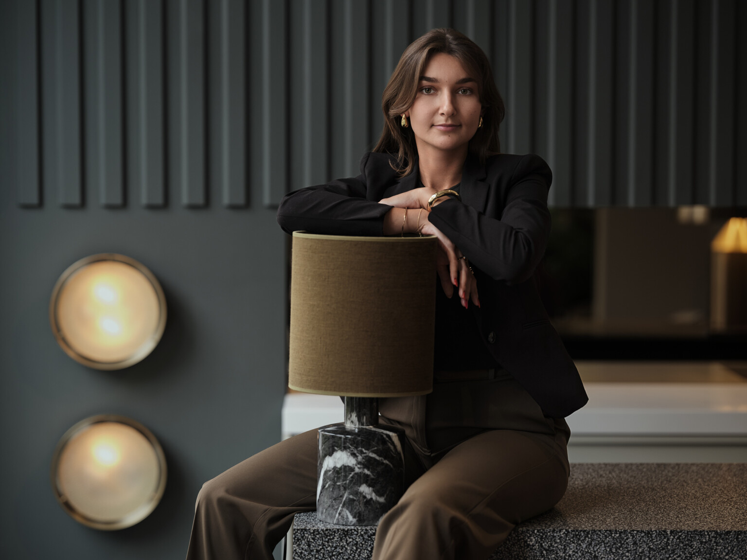 Business Portrait einer jungen Frau im schwarzen Blazer an einer Tischleuchte, dunkle Wand mit Wandleuchten
